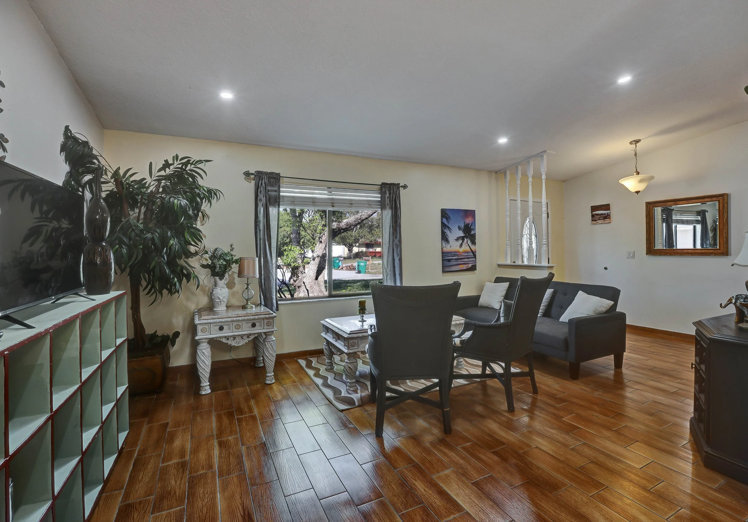 Living room with wooden floor, black chairs, gray sofa, large window with curtains, painting of sunset with palm trees, TV, small white table with decorative items, and potted plant.