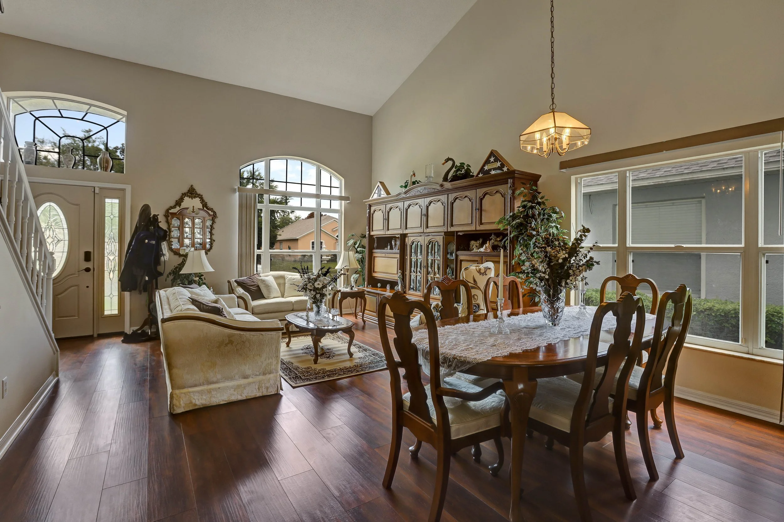 Living and dining room with wooden furniture, cream sofas, a glass-top coffee table, a wooden dining table with six chairs, a large wooden display cabinet, floral arrangements, a chandelier, and large windows letting in natural light.