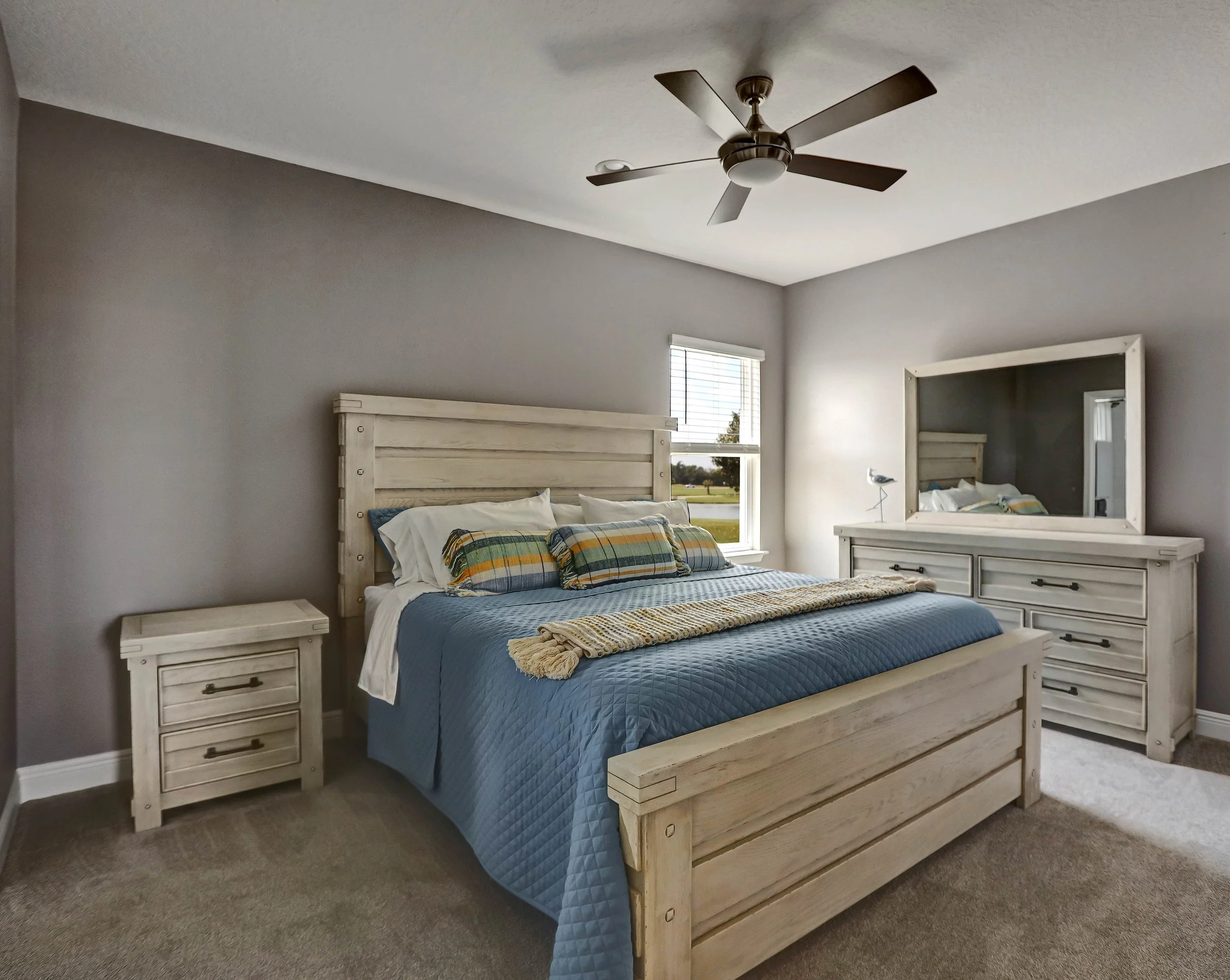 A bedroom with a white wooden bed, gray walls, a blue quilt, plaid pillows, a nightstand, a dresser with a mirror, a lamp, a window with blinds, and a ceiling fan.