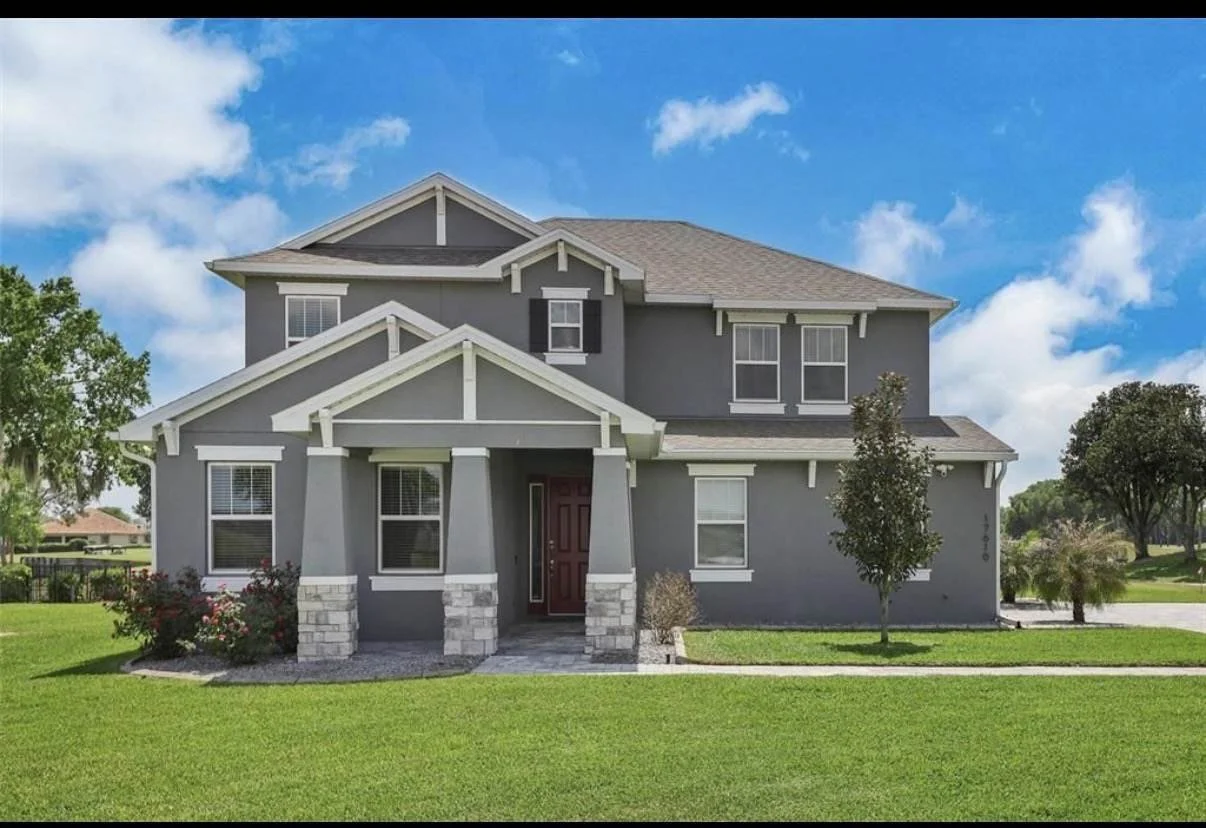 A modern two-story house painted in dark gray with white trim, featuring a front porch with stone pillars, a brown front door, multiple windows, and a well-maintained lawn with small trees and bushes. The background shows a blue sky with white clouds