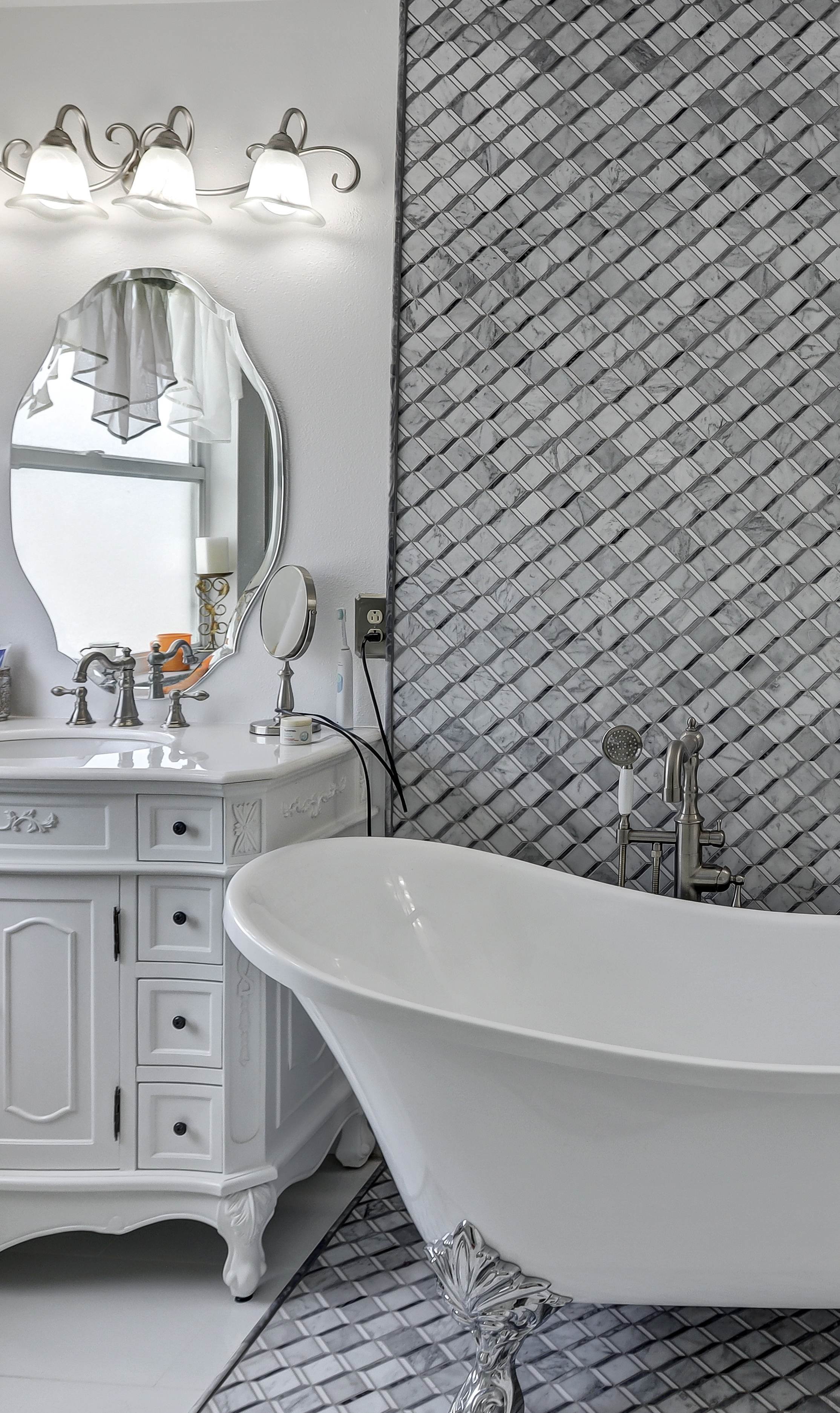 Bathroom with a vintage white vanity with a mirror, a white freestanding clawfoot bathtub, gray patterned wall tiles, and a window with a curtain.