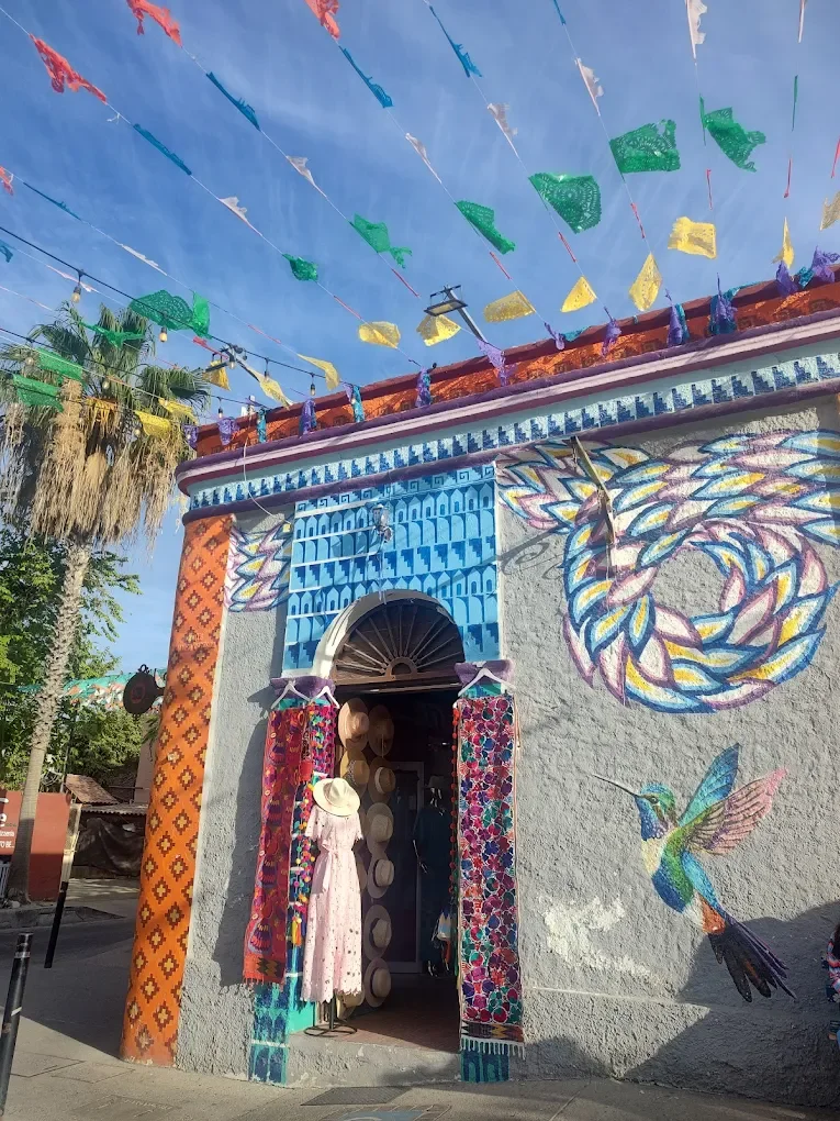 Colorful building with vibrant murals of a hummingbird and swirling patterns, decorated with textiles and hats at the entrance, with festive banners overhead and palm trees nearby in San Jose del Cabo