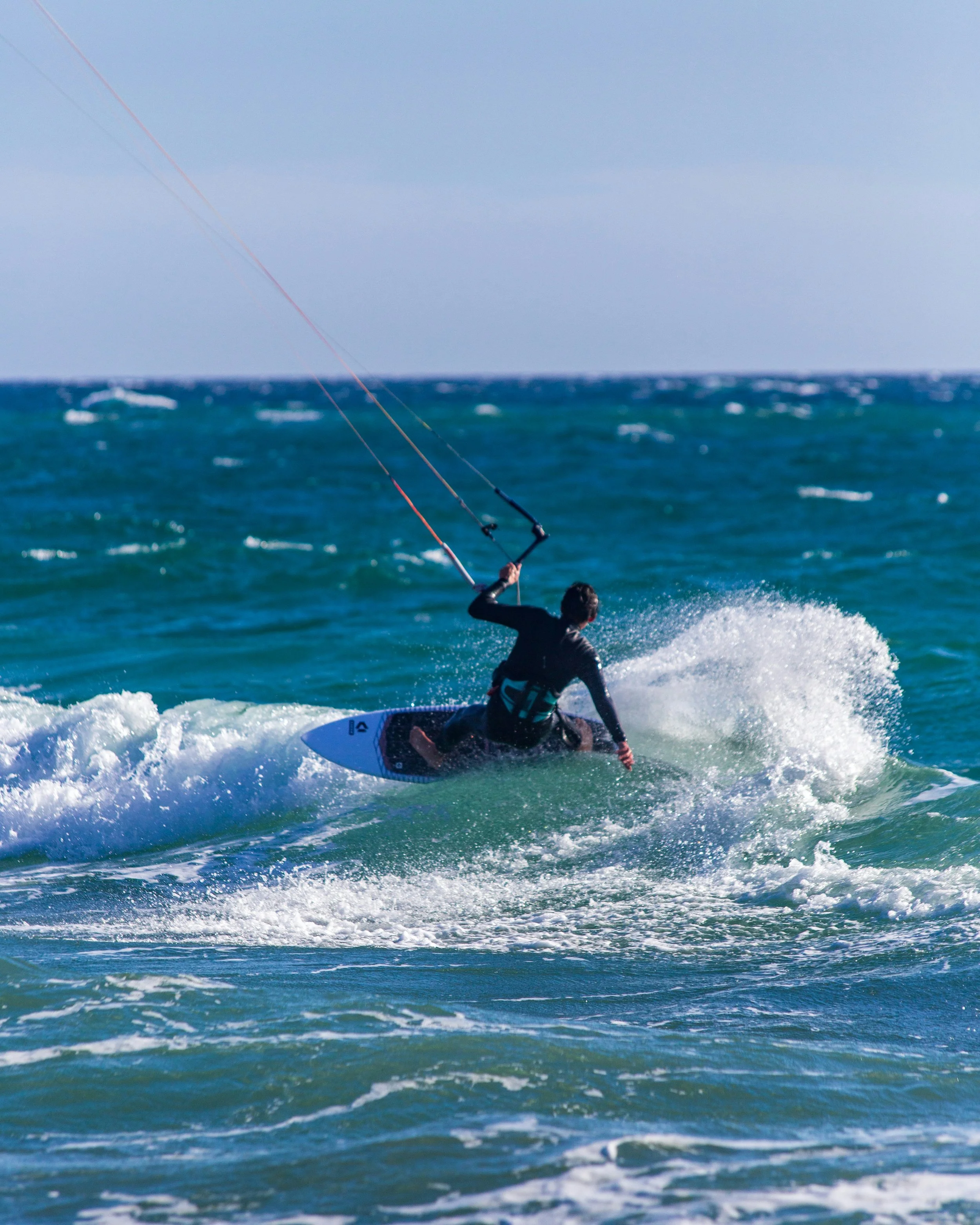 A person kiteboarding on the ocean, holding onto the control bar with long kite lines visible above the water, as waves crash around.