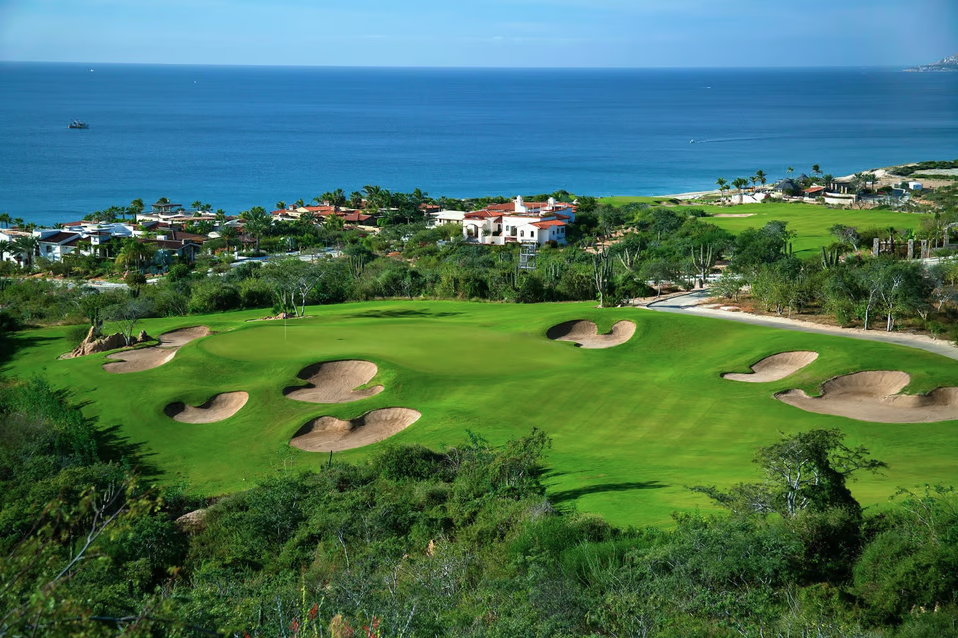 A golf course with numerous sand bunkers, overlooking the ocean with houses in the background and lush greenery in the foreground.