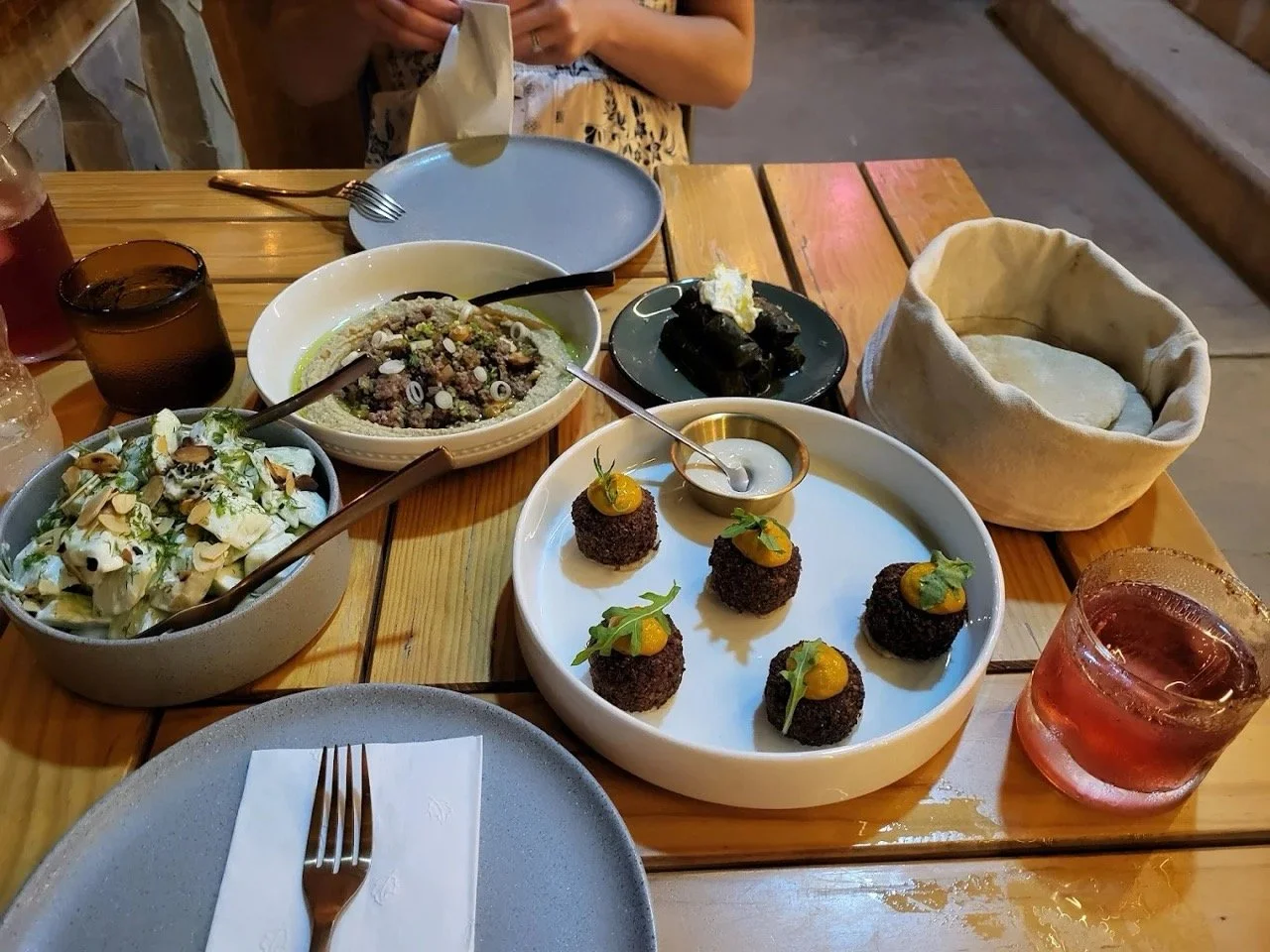 A table set with various dishes including salads, a plate of croquettes topped with sauce and greens, a small bowl of black rice or similar dish, and a black plate with a dessert, along with glasses of drinks and empty plates and utensils.