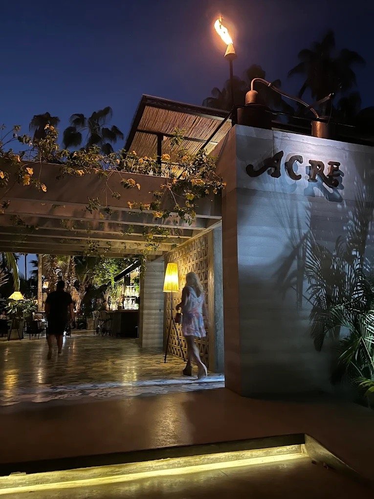 Nighttime view of the entrance to a building named 'JACRE', with decorative plants, a lit lamp, and a torch burning on top of the entrance.