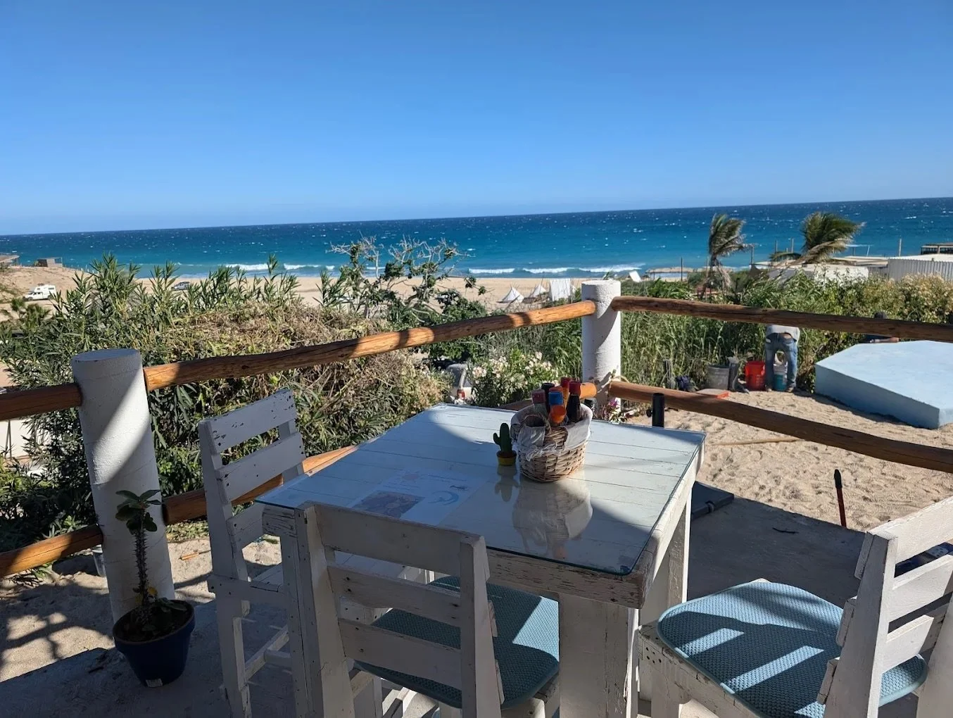 Beachside patio with white chairs and a glass table, overlooking the ocean with a sandy beach, greenery, and palm trees under a blue sky.
