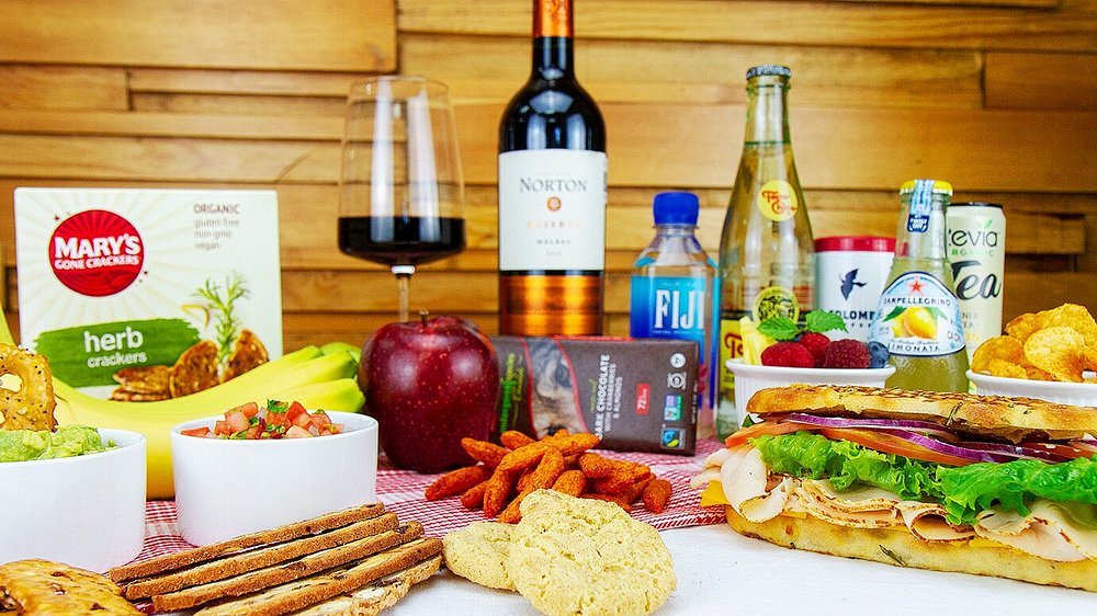 A variety of food and drinks on a table, including a sandwich, fruit, snacks, wine, and bottled beverages, with a wooden wall background.