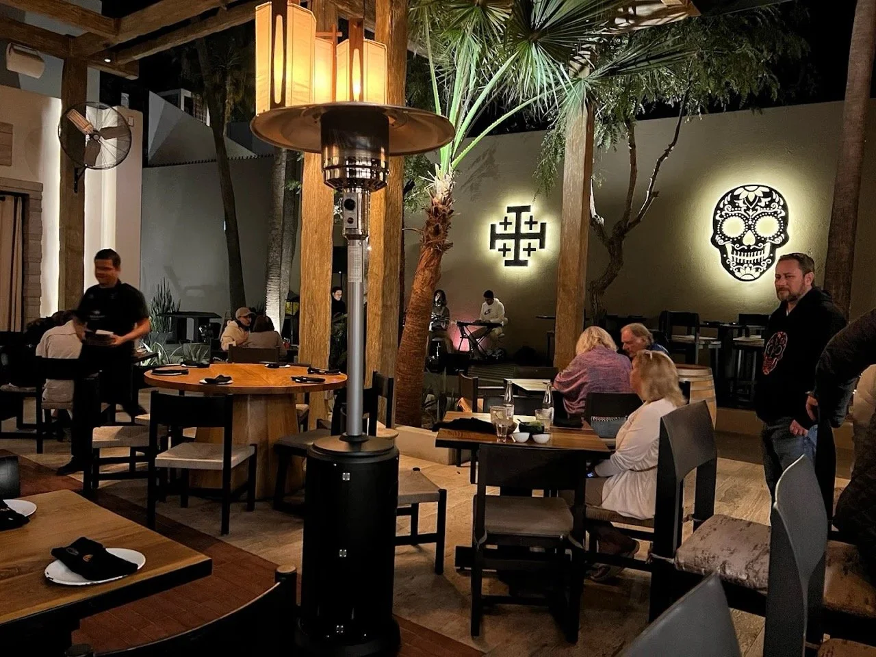 Indoor restaurant with wooden tables, chairs, and a large patio heater. Decor includes illuminated symbols and a sugar skull wall art, with several patrons dining and staff serving.