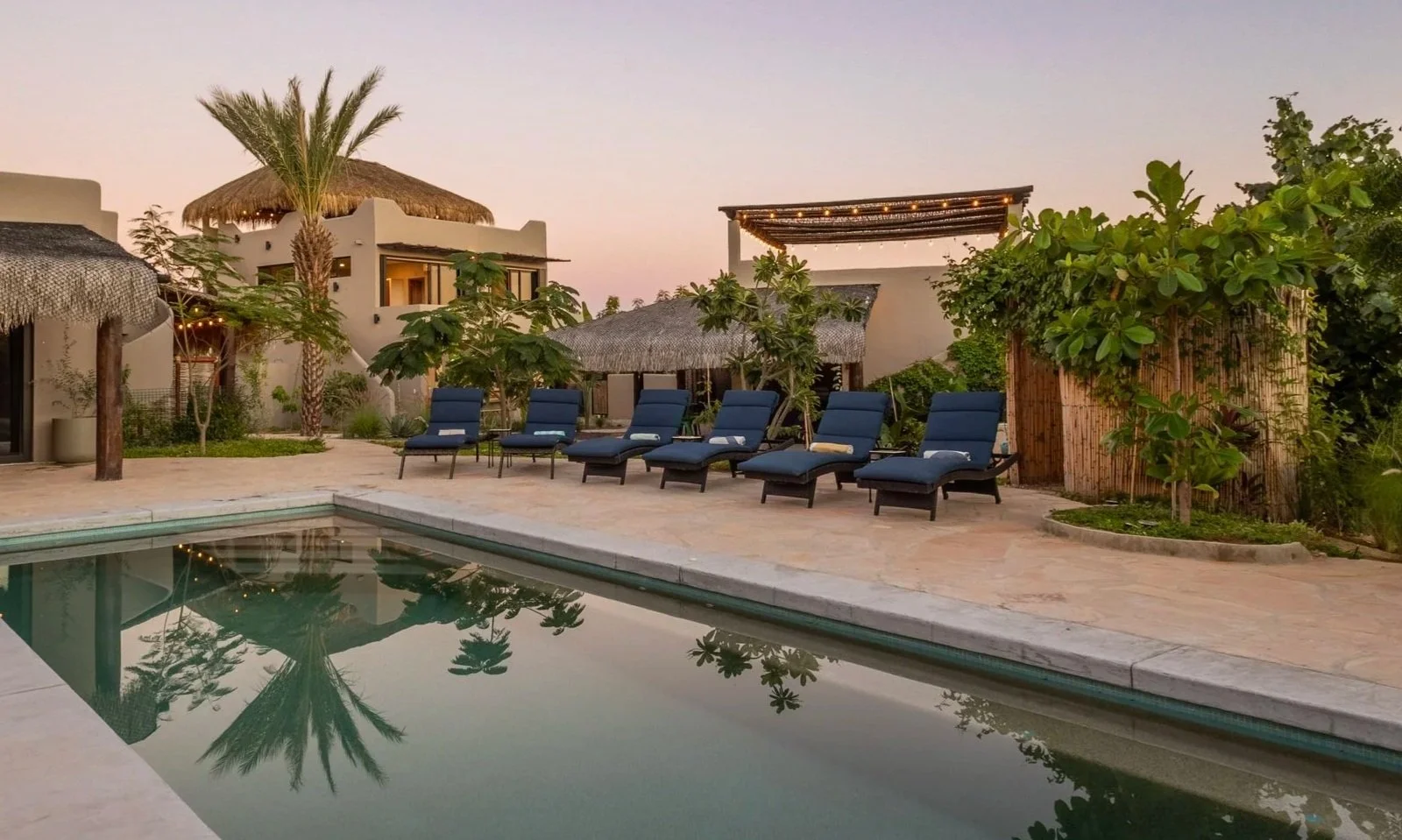 Poolside scene at sunset with blue lounge chairs, surrounded by palm trees and thatched-roof buildings, reflecting in the calm water.