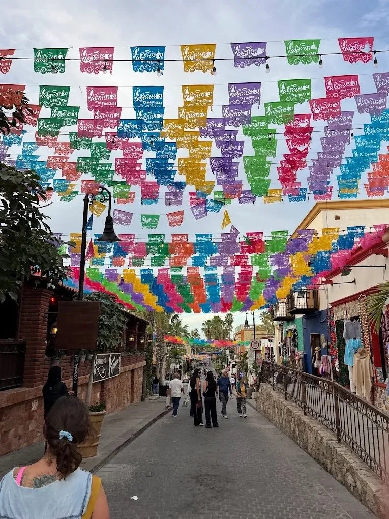 Colorful papel picado banners hanging across a busy street San Jose del Cabo during a festive day, with people walking and shops on the sides.