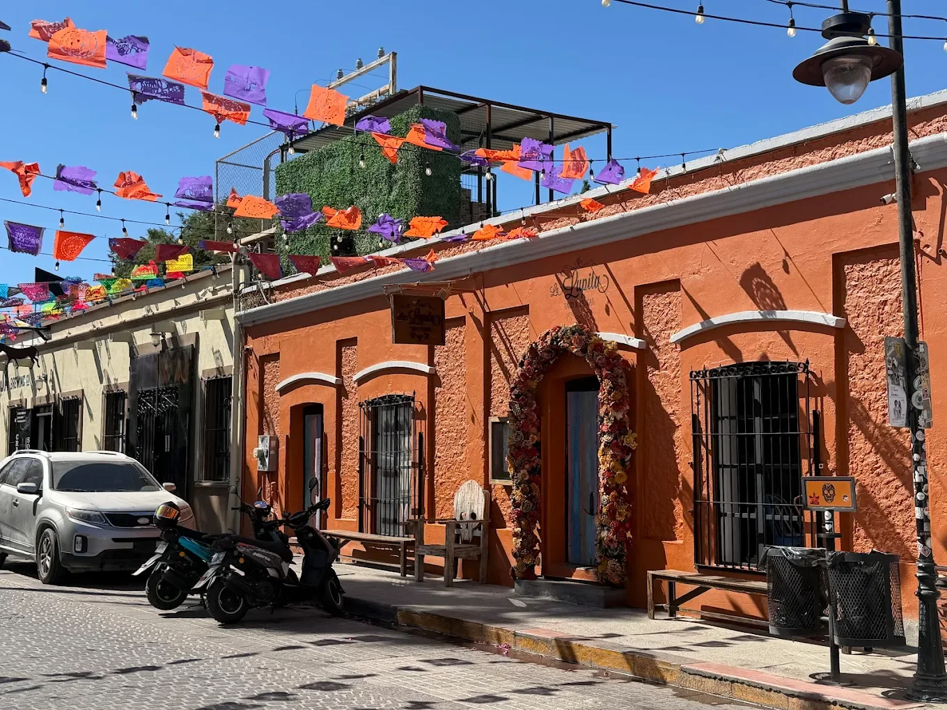 Street view of colorful orange and beige buildings with decorated entrance, parked car and motorcycle, bright sunny day, festive paper banners hanging overhead.