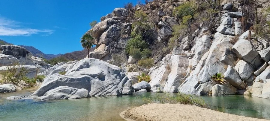 A desert landscape with large gray rocks surrounding a small river or creek, with sparse vegetation and a bright blue sky.