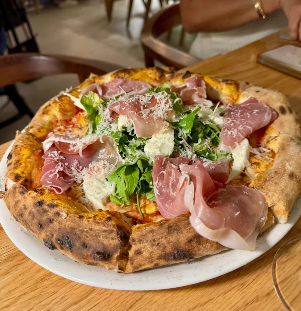 Pizza with prosciutto, fresh greens, and shredded cheese on a wood table.