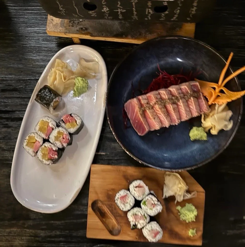 Sushi rolls, sashimi, and raw fish on plates and a black plate, with wasabi, ginger, and vegetables, on a black wooden table.