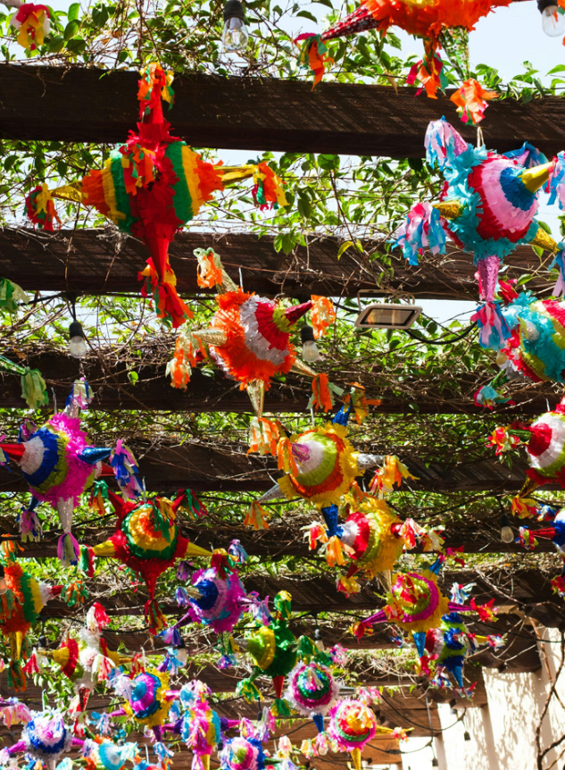 Colorful piñatas hanging from a wooden pergola among green vines and string lights.