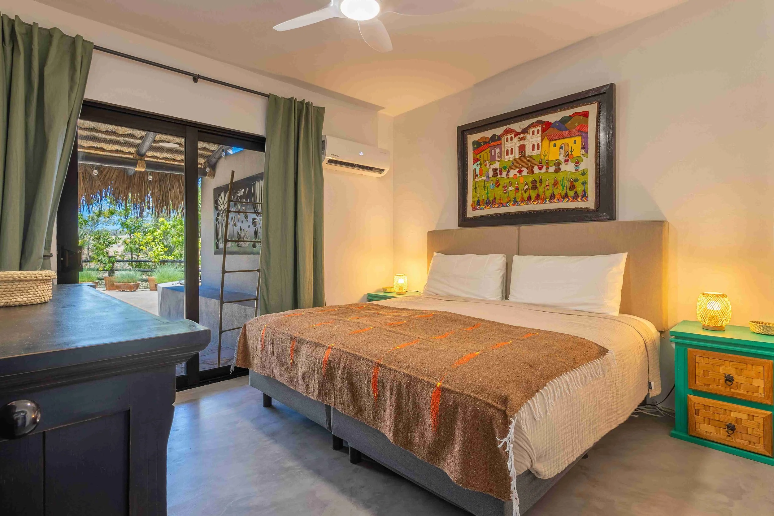 A cozy bedroom with a large bed, green nightstands, and a painting above the bed. There are green curtains covering a sliding door leading to a balcony with lush greenery outside.