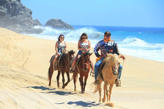 Three people riding horses along a sandy beach with the ocean and cliffs in the background.