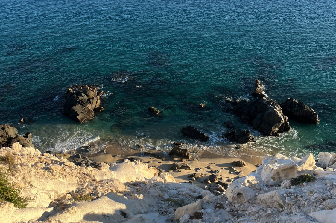 A rocky beach with clear blue-green water and large rocks in the ocean, viewed from a sandy cliff.