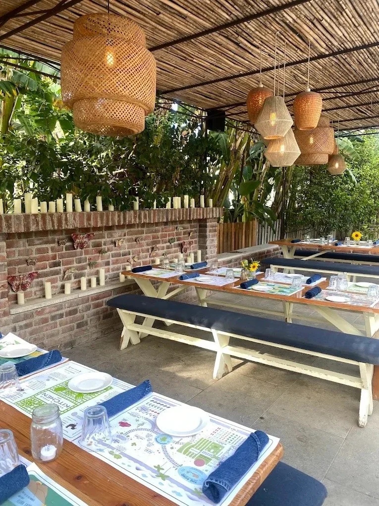 Outdoor restaurant patio with wooden tables set with plates, glasses, and napkins. Blue napkins are rolled on each table. Decor includes sunflower centerpieces, a brick wall with candles, and wicker pendant lights hanging from a bamboo ceiling. Green plants and trees surround the area.