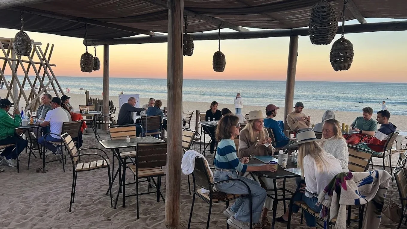 Beachside restaurant with groups of people dining at tables during sunset, with the ocean and sky in the background.