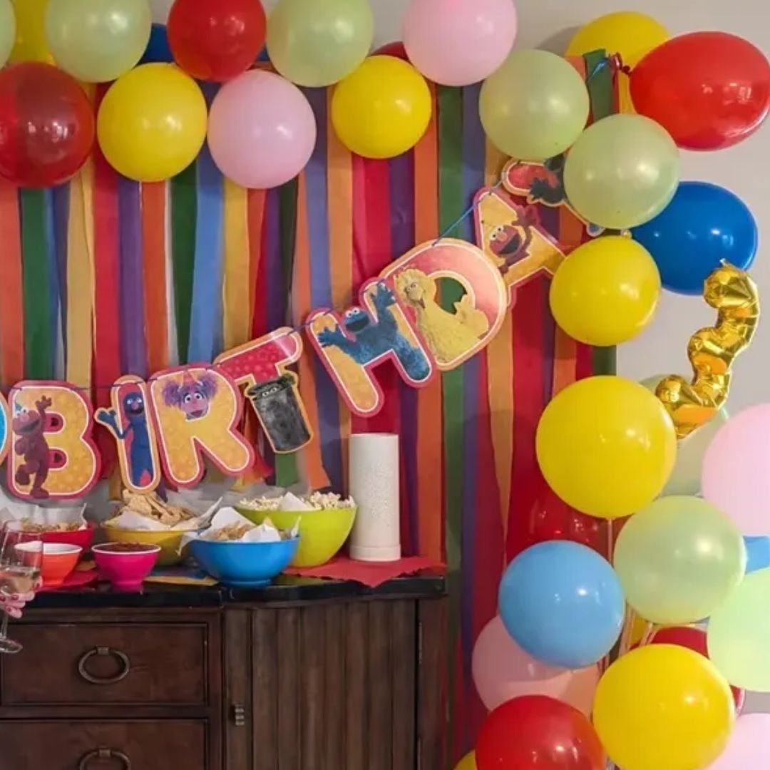 Birthday party decorations with multicolored balloons and a Sesame Street themed banner. Streamers and bowls of snacks are on a table.