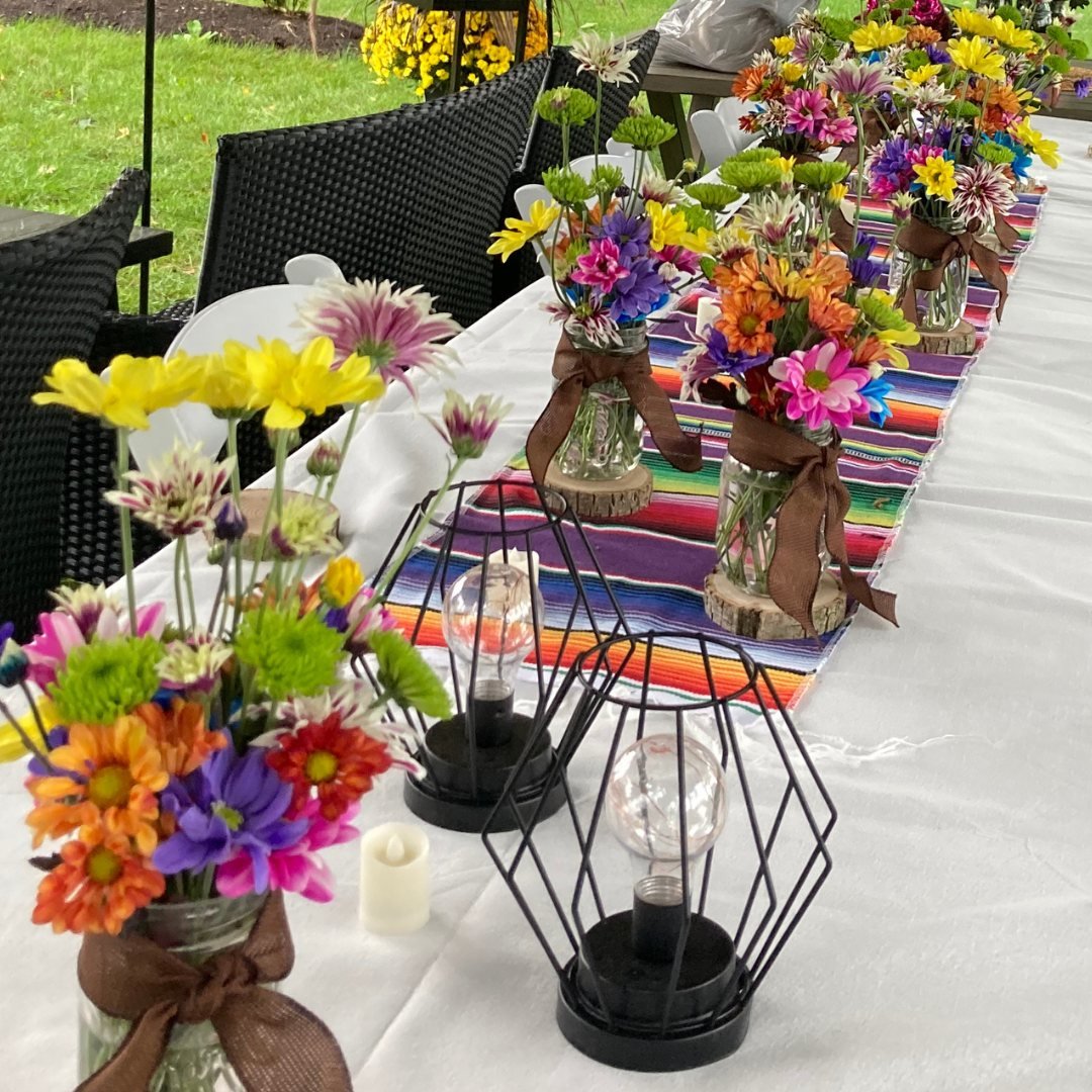 A outdoor table setup with colorful floral centerpieces and decorative elements. The table features a white tablecloth, vibrant flowers in jars tied with brown ribbons, black wire cages as table decor, and a multicolored striped runner. The setting s