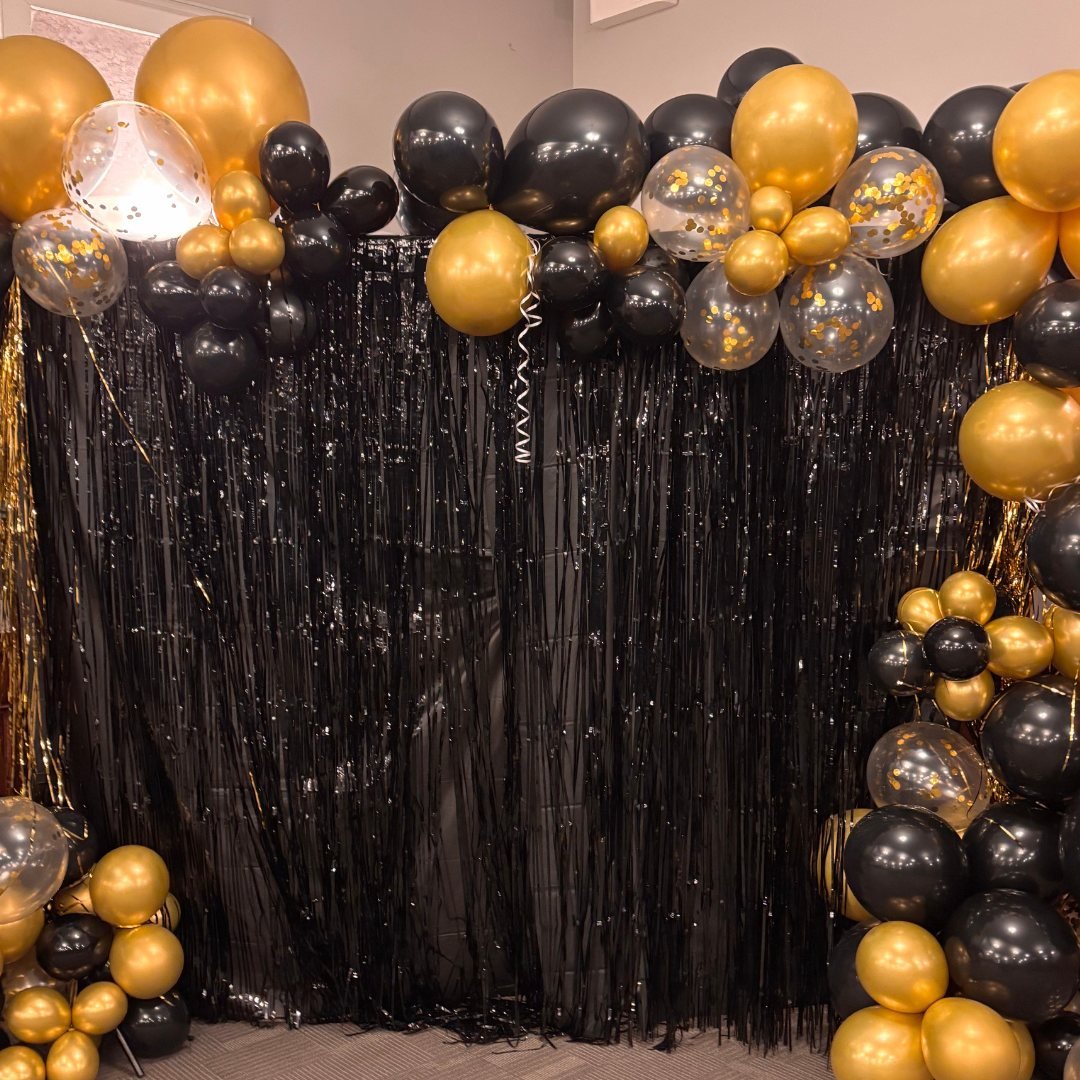 Black and gold party balloon arch with black fringe backdrop.