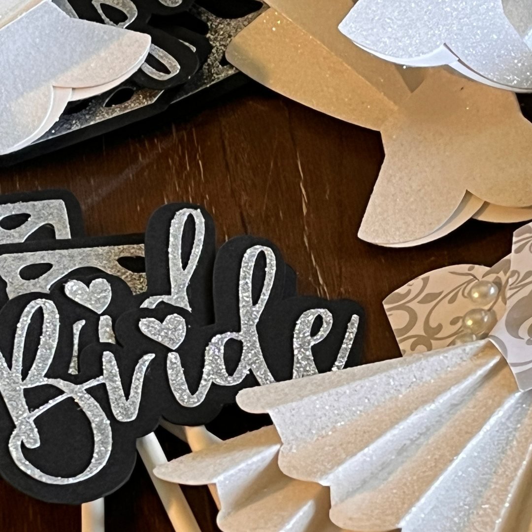 Decorative cutouts for a bridal shower, featuring black and silver glitter "Bride" text and white paper dress designs.