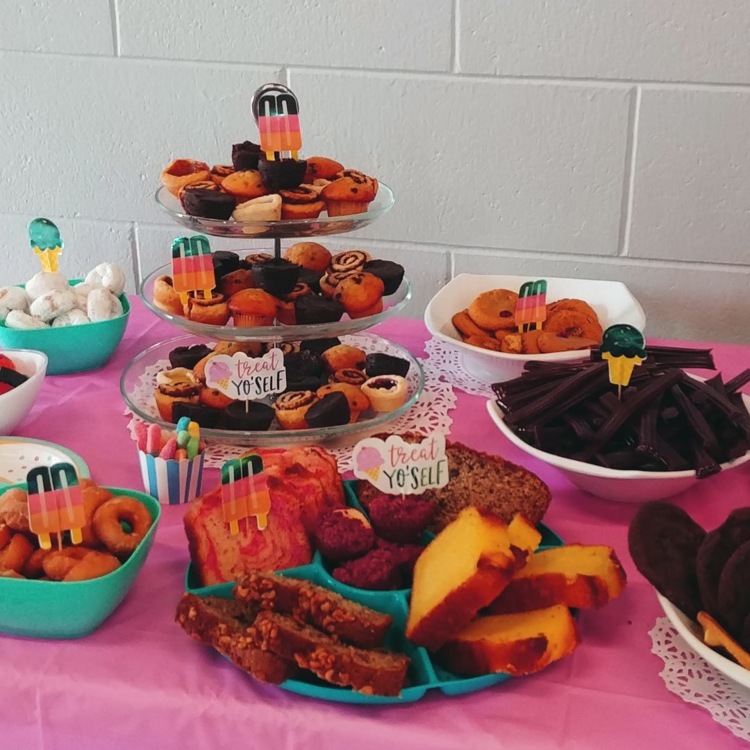 A dessert table with various treats including a tiered stand of muffins and pastries, bowls of cookies, licorice, and donuts, and plates of assorted breads. Decorative signs read "Treat Yo' Self."