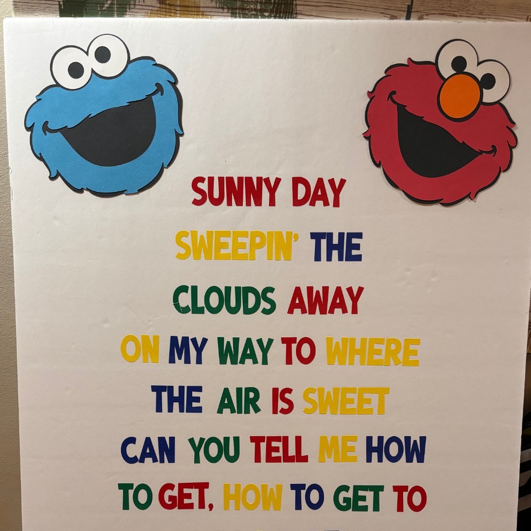 Poster with colorful text displaying lyrics from a children's song, featuring cartoon faces of a blue and a red character.