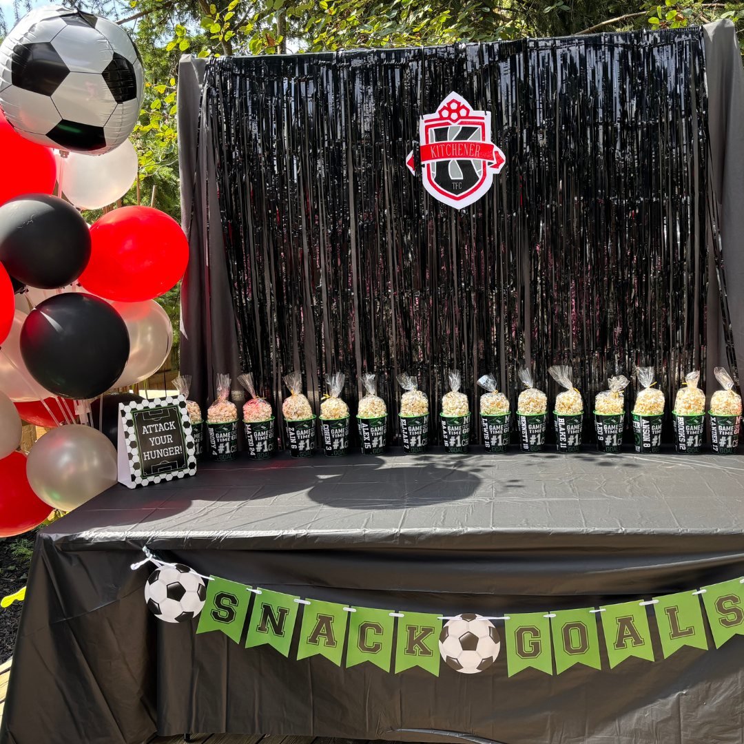 Soccer-themed snack table with a black backdrop, popcorn bags, balloons, and decorative signage.
