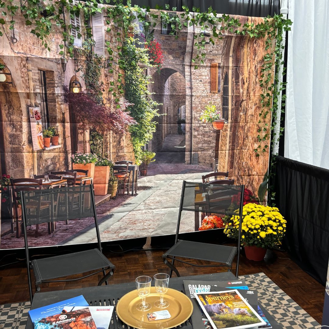A cozy booth setup with a backdrop depicting a scenic European alleyway, featuring stone buildings and plants. In front, there are two empty chairs, a table with magazines and brochures, two empty glasses on a golden tray, and a pot of yellow flowers
