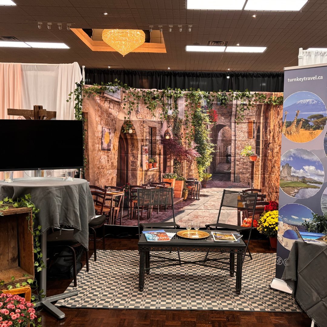 Travel expo booth with Mediterranean street backdrop, chairs, a table with brochures, and a TV screen.
