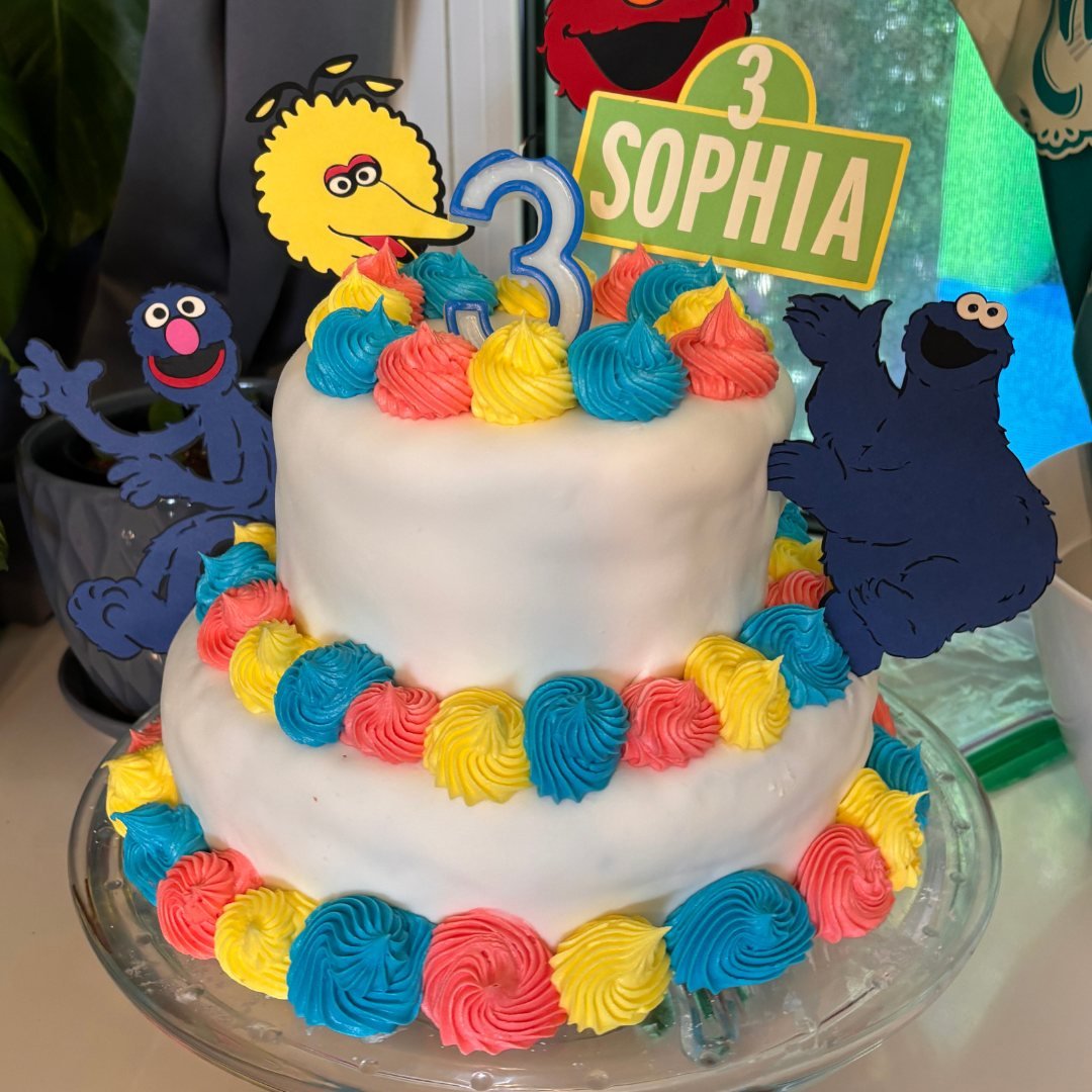 Two-tiered birthday cake with white icing and colorful frosting decorations in blue, yellow, and pink. Topped with cutouts of characters resembling those from a popular children's show, along with a number '3' and a sign reading 'Sophia.'
