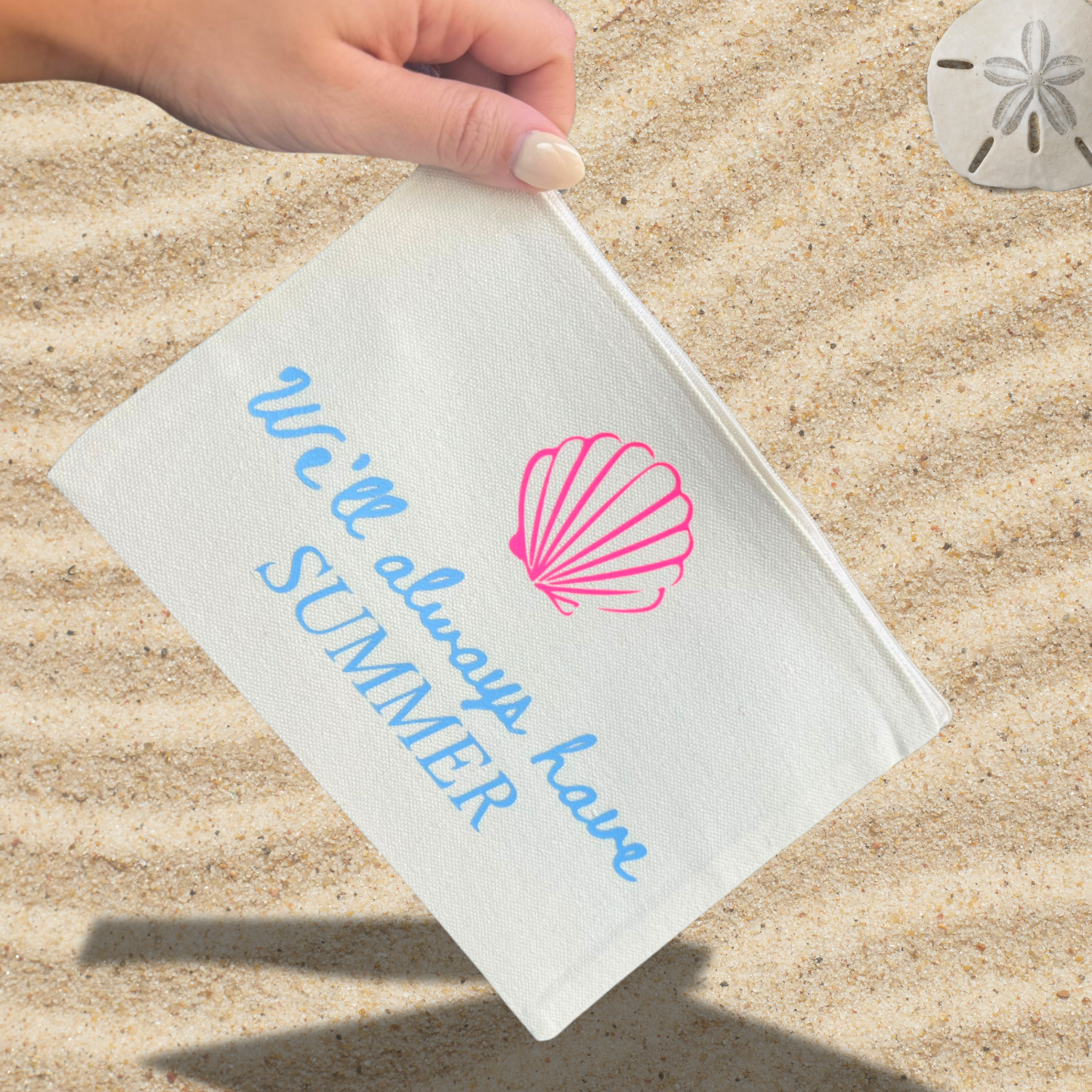 A hand holding a white pouch with text "We'll always have SUMMER" and a pink shell illustration, placed on a sandy beach background with a sand dollar.
