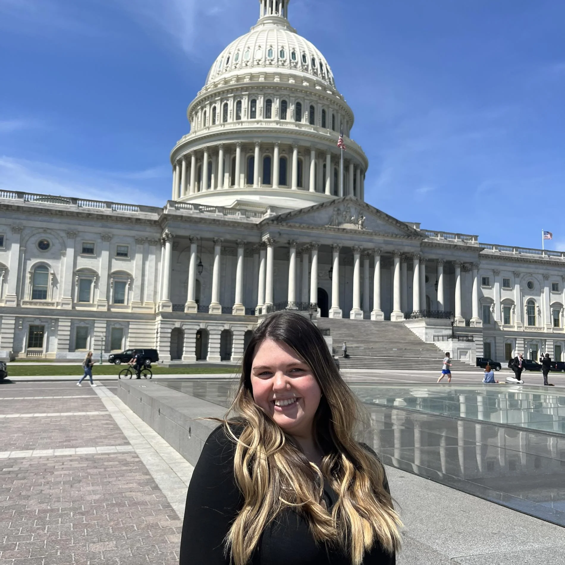 Smiling woman with long blonde hair standing in front of the United States Capitol building on a sunny day.