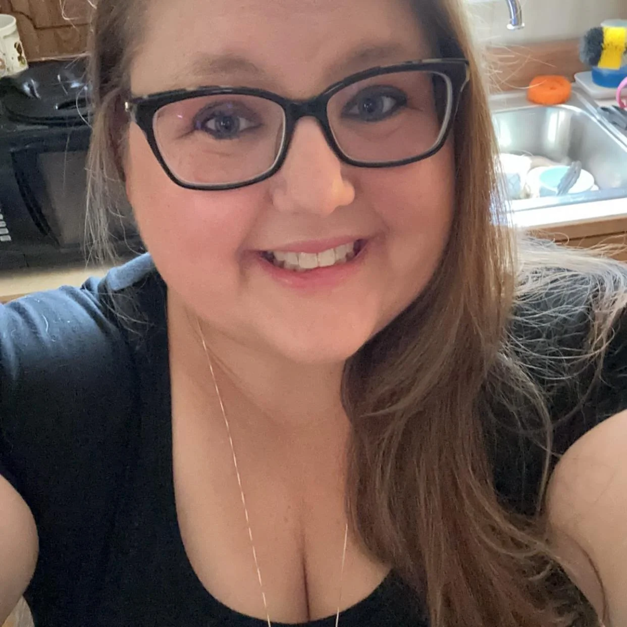 A woman with long brown hair and glasses smiling in a kitchen.