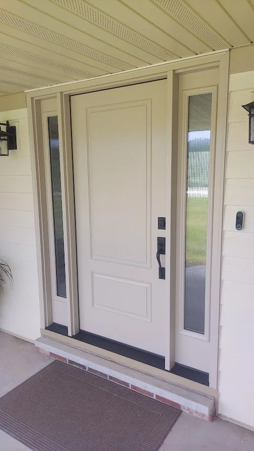Front door with sidelight windows on either side, white door with black handle and lock, brick step, brown doormat, wall-mounted lamp and doorbell