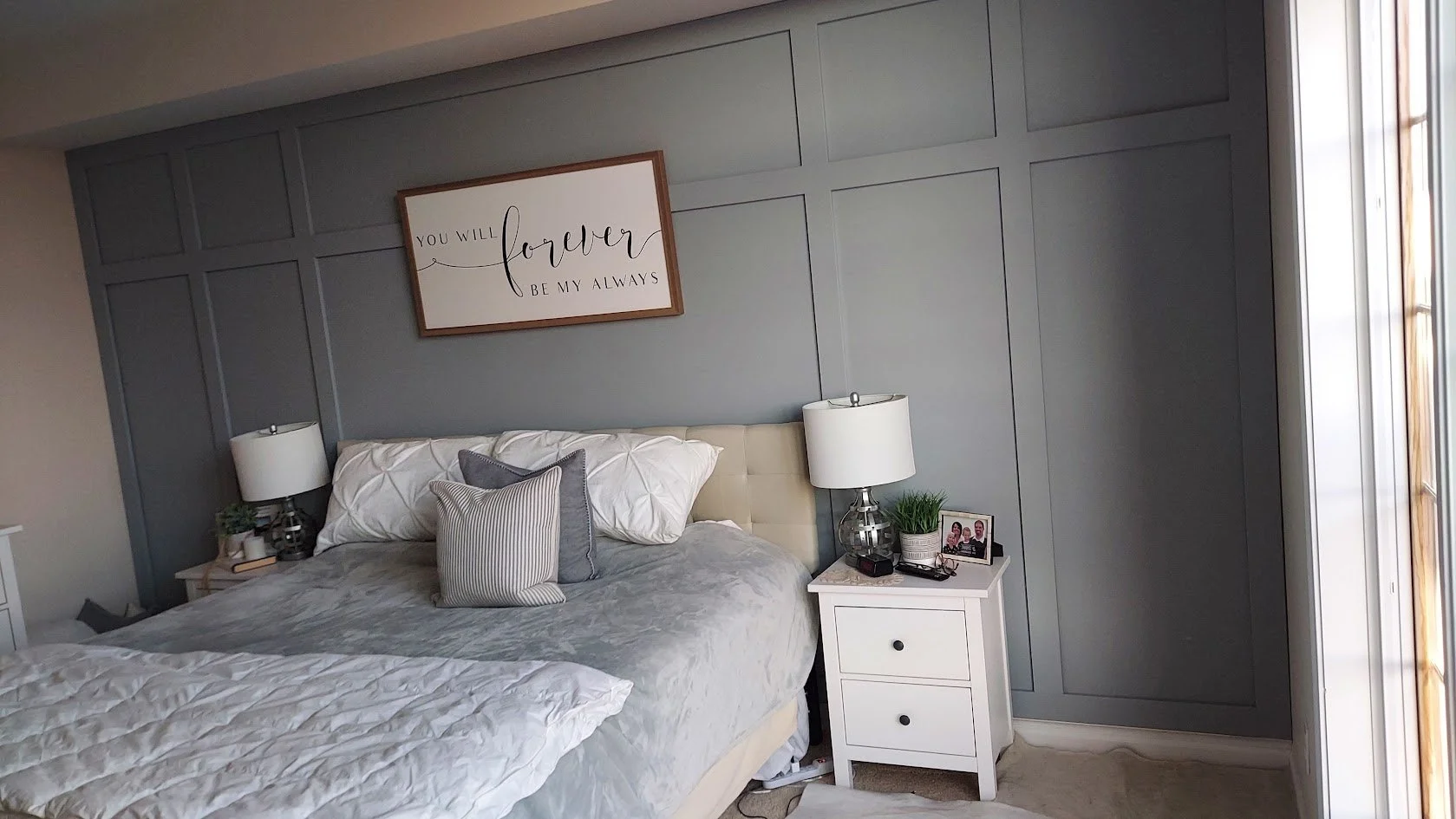 A bedroom with a gray paneled wall, a white upholstered bed with gray and white pillows, and a white nightstand with a lamp, a small plant, and framed photos.