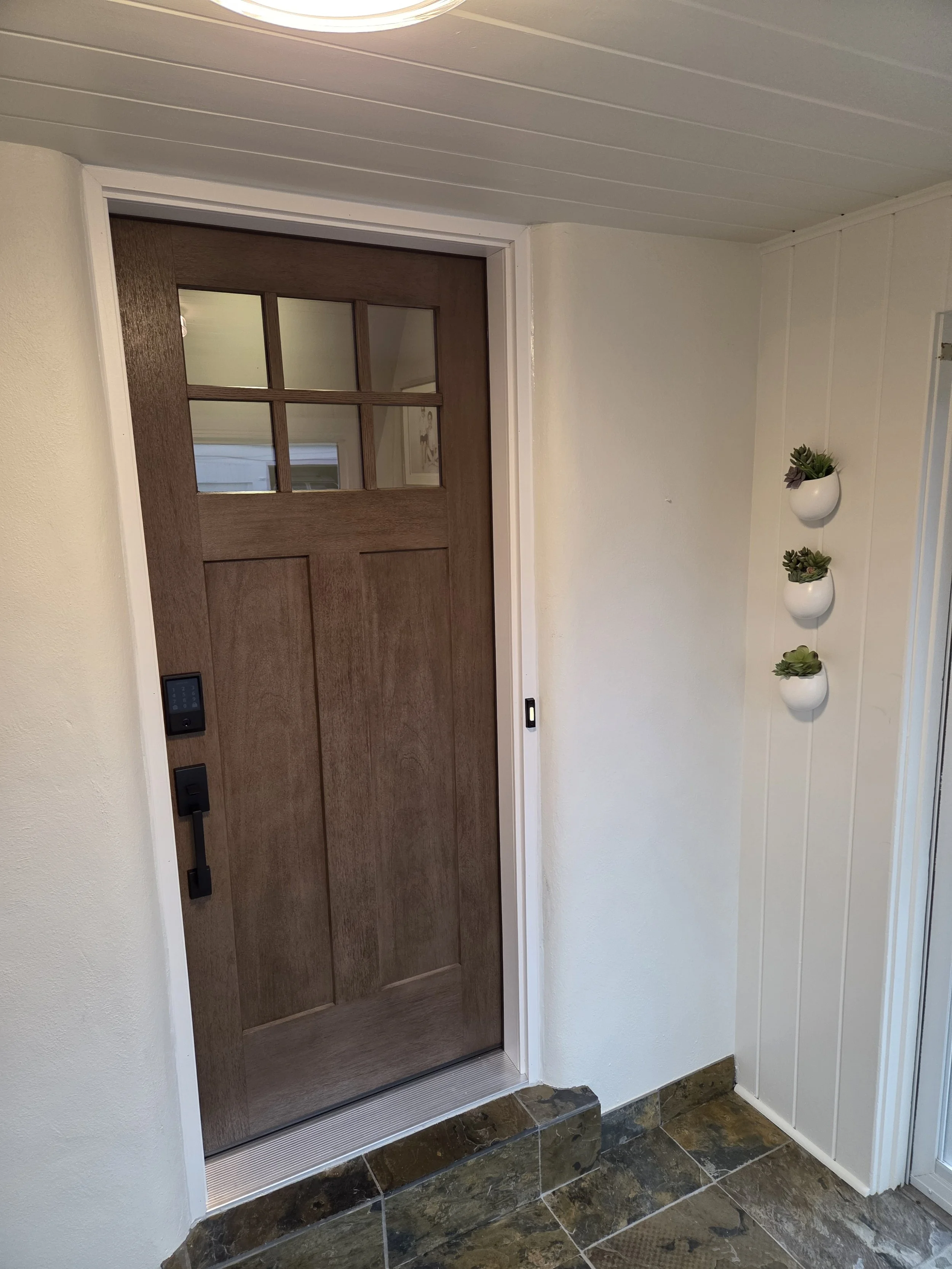 A wooden front door with a small window at the top, set in an entryway with white walls and a tile floor. There are three small white planters with succulents hanging on the wall to the right of the door.