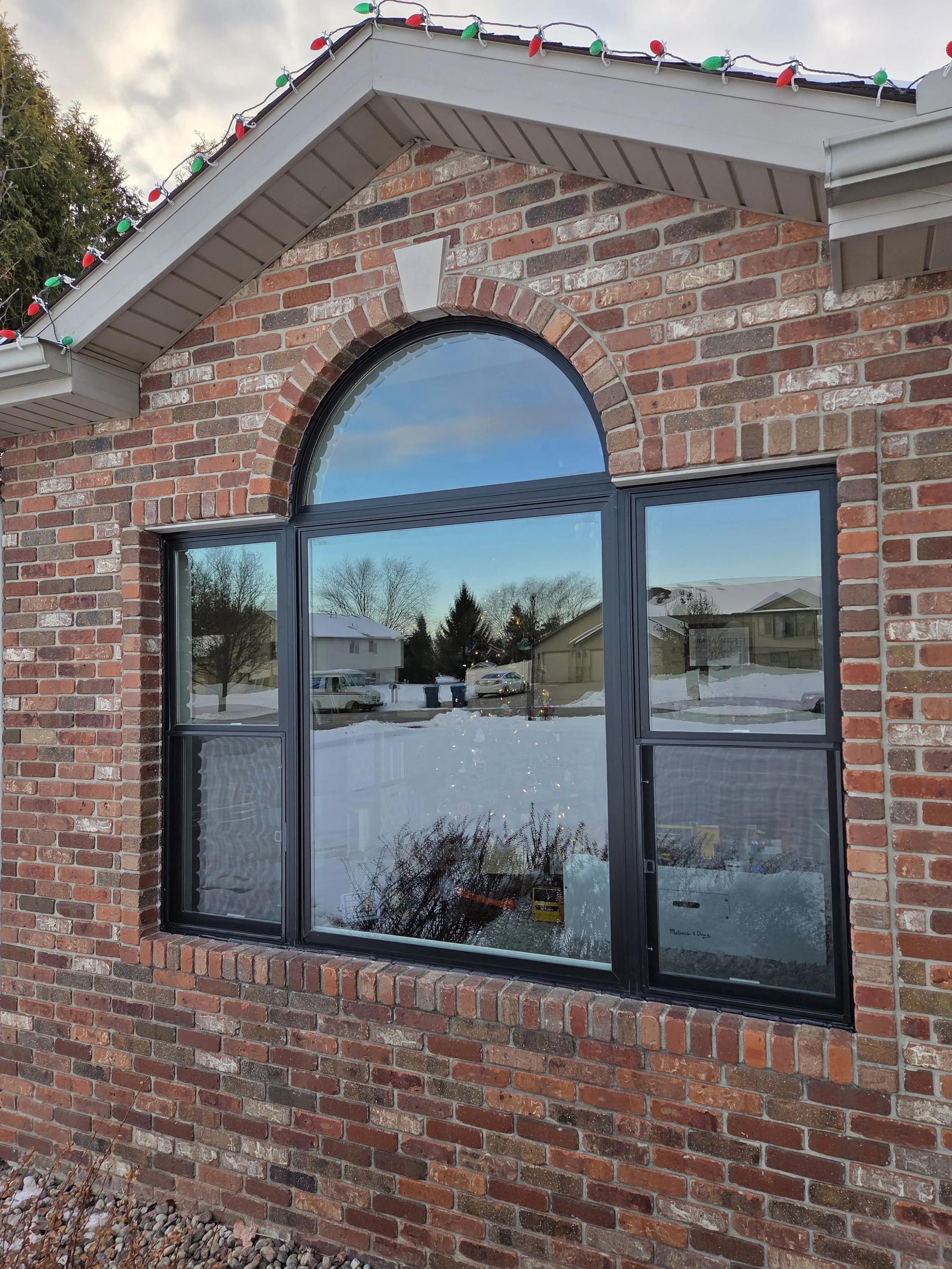 A brick house featuring a large, arched window with black framing, reflecting a snowy suburban neighborhood with houses, trees, and parked cars. The eaves are decorated with colorful Christmas lights.