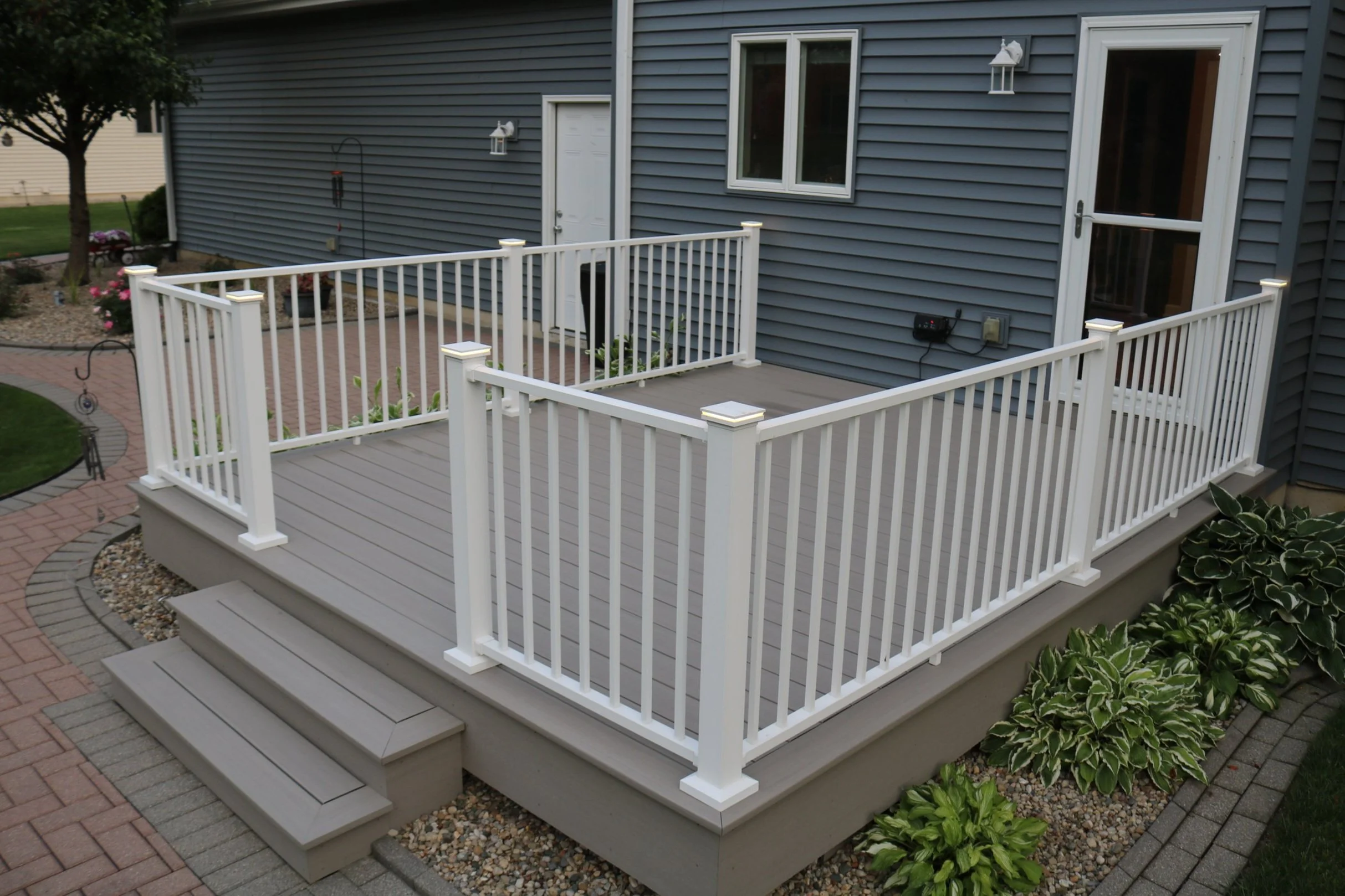 A backyard deck with white railing, gray flooring, and steps leading down to a brick-paved walkway, surrounded by landscaped plants and garden beds.