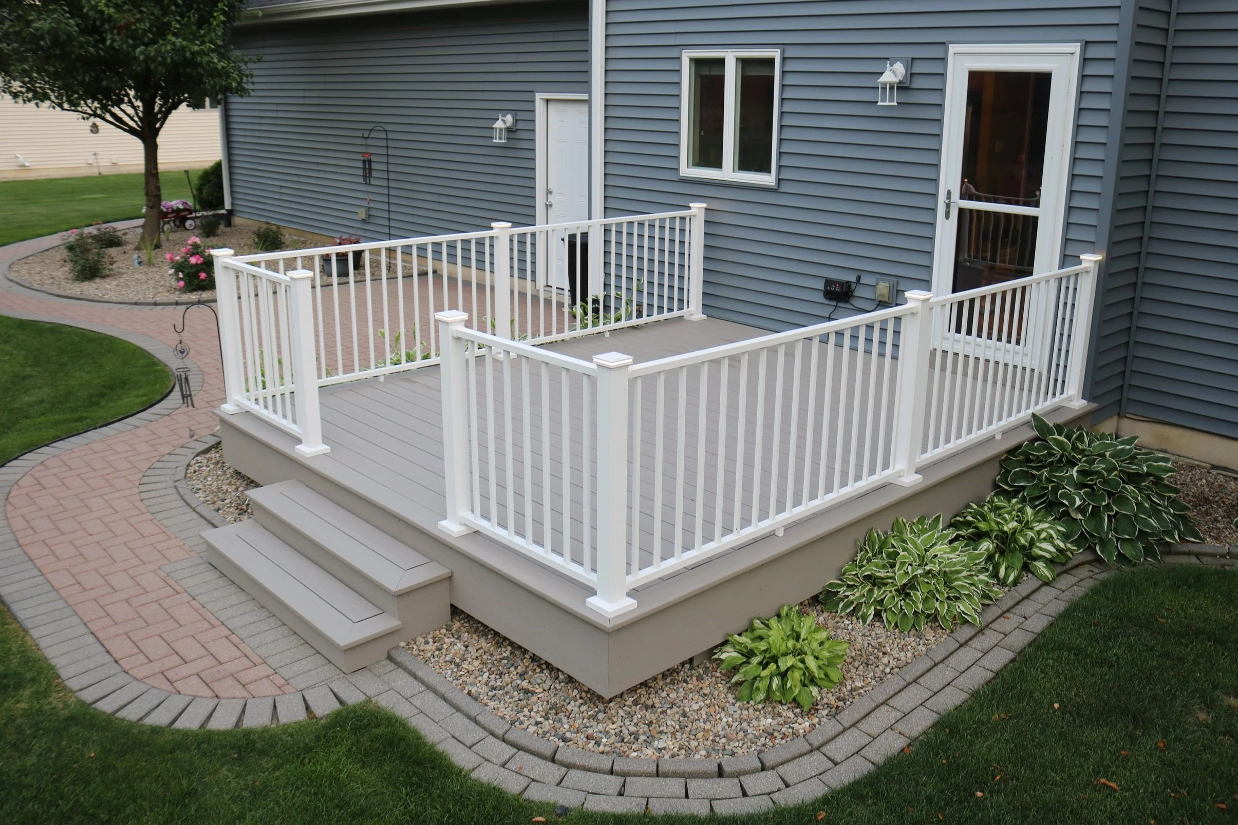 Backyard with a gray deck, white railings, steps, landscaped plants, and a curved brick pathway leading to a house with gray siding.