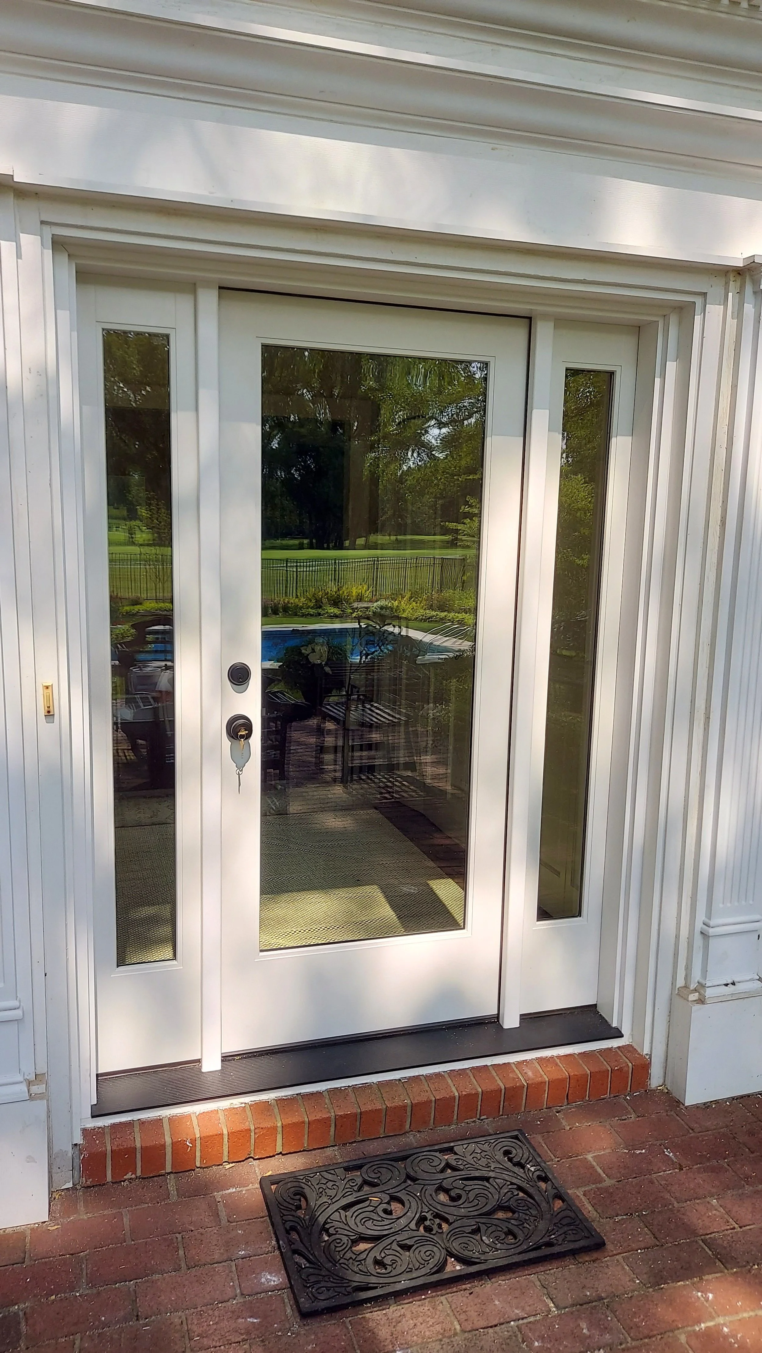 Exterior view of a glass door with sidelights, showing reflections of a backyard with a pool and trees.