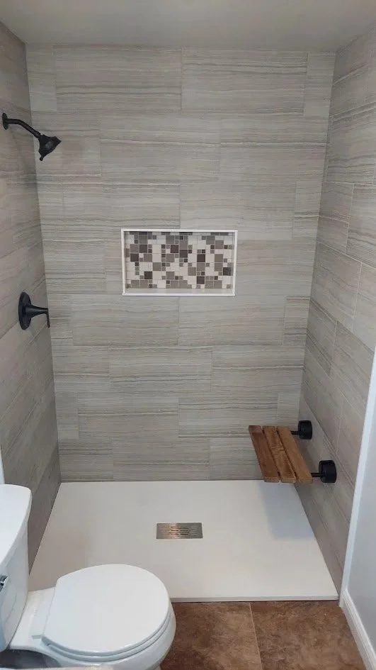 Custom walk-in shower with beige tiling, a small mosaic tile niche, a wooden shower bench, and a toilet in a bathroom.