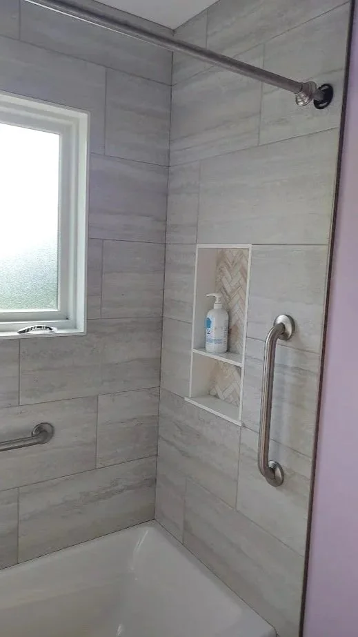 Custom tiled shower with a window, two metal grab bars, a built-in shelf with a bottle, and a shower curtain rod.