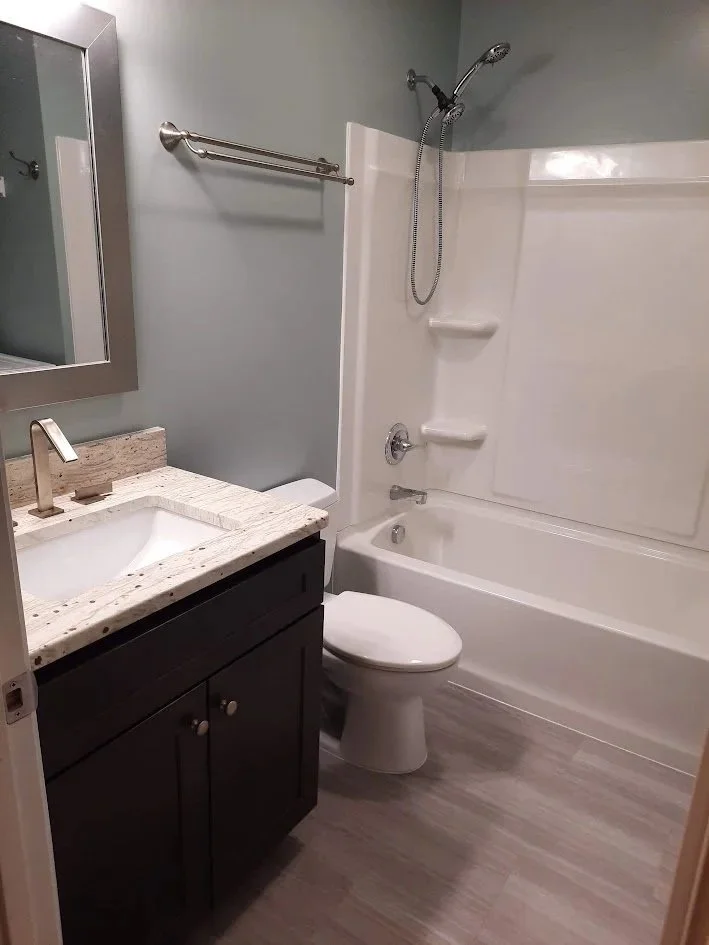 Small bathroom with a white bathtub and shower, toilet, dark vanity with a sink, and a mirror. Light gray wood-like flooring and pale blue walls.