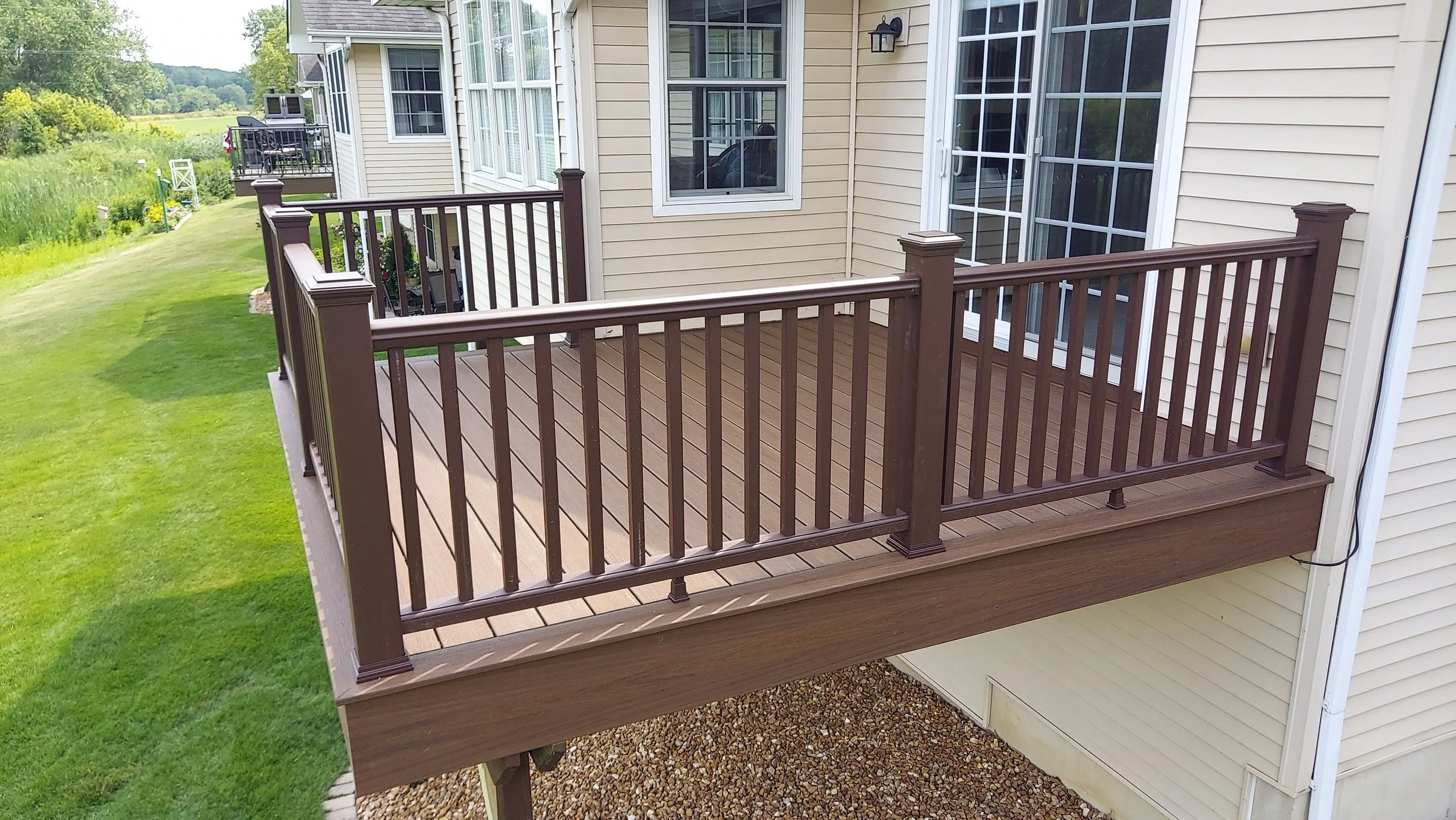 A wooden balcony attached to a beige house with beige siding, featuring brown railings and matching posts. The balcony is elevated above a gravel ground area and overlooks a well-maintained green lawn and scenic landscape.