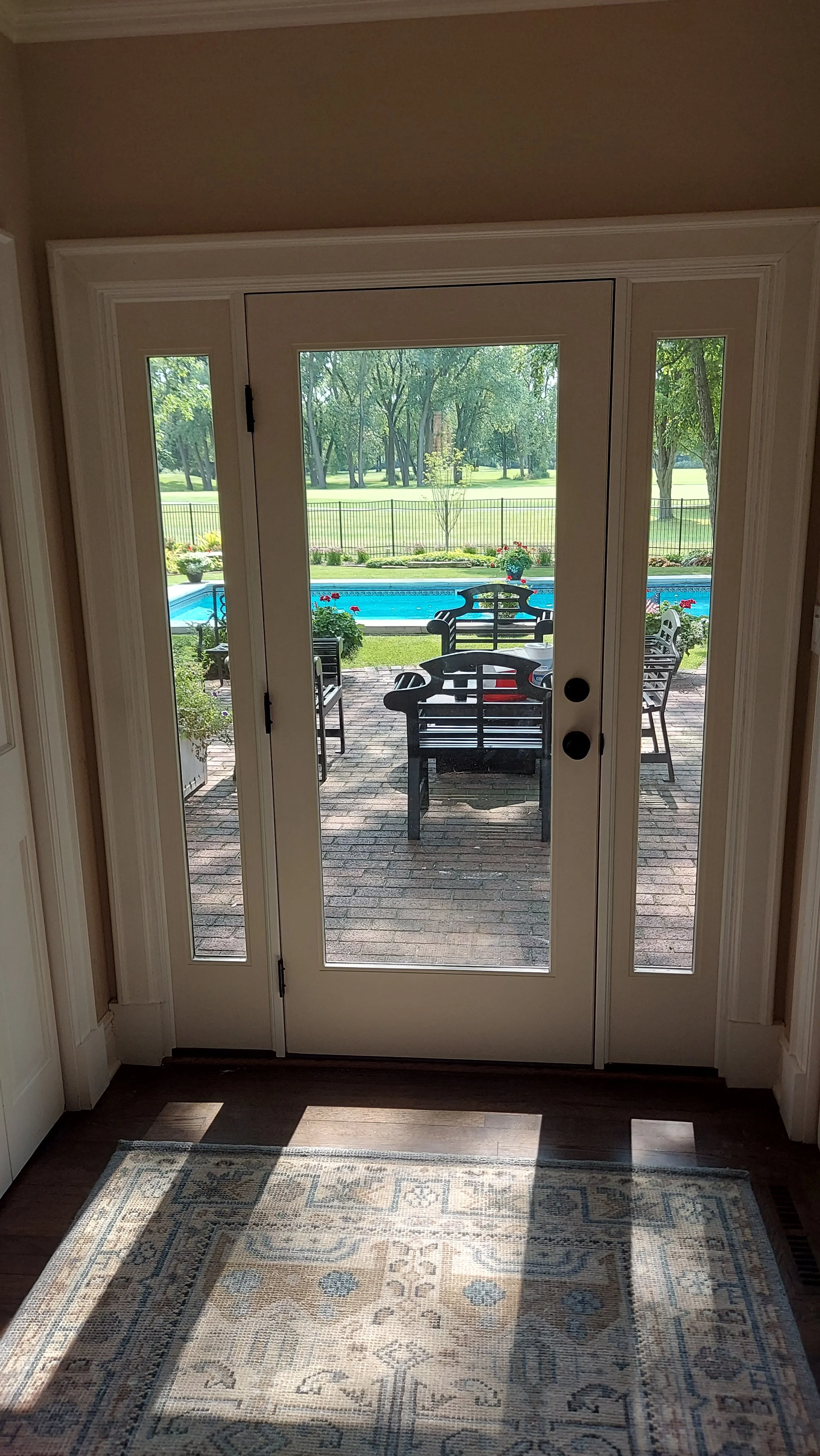 View through glass door showing outdoor patio with benches, a swimming pool, and a grassy yard with trees.
